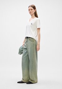 Biely tričko s V výstrihom a logom CK, kombinované so širokými, vyblednutými zelenými džínami. Model drží svetlomodrú bundu a má na sebe čierne topánky.