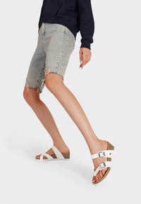 Sandali bianchi senza chiusura con cinturini incrociati e soletta in sughero. La suola è strutturata, con un leggero design a zeppa per un'altezza aggiuntiva.