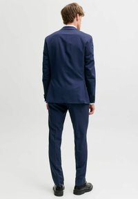 Uomo che indossa un completo aderente blu navy con scarpe eleganti nere, di spalle contro uno sfondo bianco semplice.