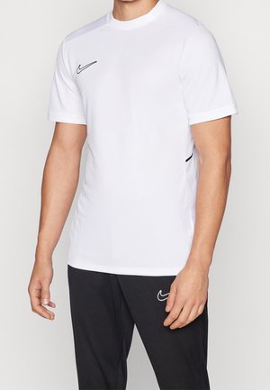 Homme portant un t-shirt Nike blanc uni à manches courtes et un pantalon Nike noir, debout devant un fond neutre.