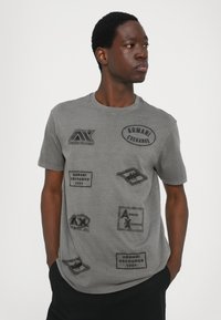 Hallhalli hallide T-särk, millel on mitmeid musti tikitud pahklappe, millel on "Armani Exchange" bränd ja erinevad kujundid ning mustrid.