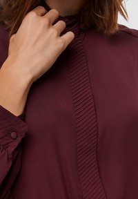 Maroon blouse with a pleated front panel, buttoned cuffs, and a high neckline, made of lightweight fabric with a smooth texture.