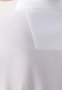 Gros plan du haut du dos d'une personne portant un polo blanc texturé avec une couture verticale près de l'épaule.