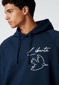 Sweat à capuche bleu marine avec un cordon de serrage, arborant une broderie blanche d'un oiseau et le mot "liberté". Texture douce et coupe décontractée.