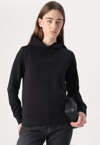 Sweat à capuche noir en tissu doux avec des manches et un ourlet côtelés. Présente un logo dans une teinte légèrement plus foncée. Associé à une pochette noire.