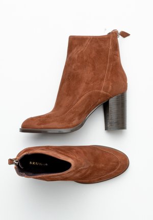 Botas de tobillo de ante marrón con puntera redonda, cremallera lateral y un tacón bloque texturizado en marrón oscuro. La parte superior presenta un detalle de costura.