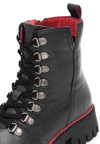 Černá kožená kotníková bota s červeným podšívem, která má kovové očko, černé tkaničky a silnou, texturovanou podrážku s červeným akcentem.