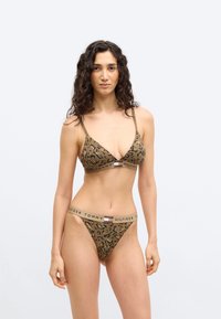 Γυναίκα με μακριά σγουρά μαλλιά, ντυμένη με καφέ σουτιέν Tommy Hilfiger με μοτίβο λεοπάρ και αντίστοιχα κοντά σλιπ, στέκεται μπροστά σε απλό ανοιχτόχρωμο φόντο.