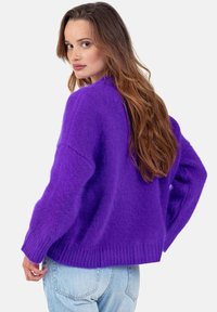 Pull en maille violet vibrant avec une coupe décontractée, des poignets et un ourlet côtelés, et une texture douce et duveteuse. Porté avec un jean bleu clair.