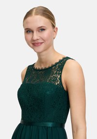 Dark green lace dress with a fitted bodice, sheer lace neckline, and a flowing skirt. Features a satin belt and floral lace pattern.