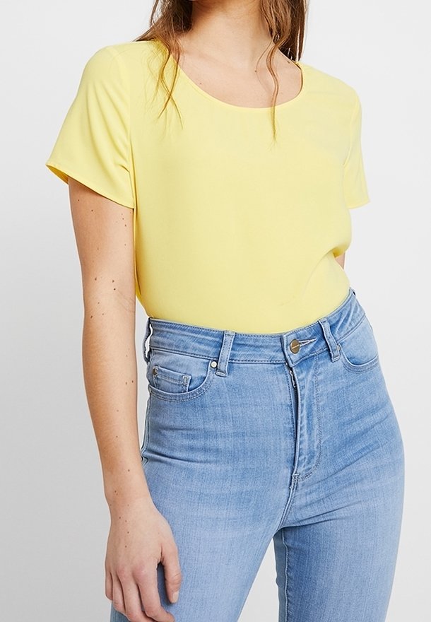 Femme portant un haut jaune à manches courtes rentré dans un jean bleu clair taille haute, debout devant un fond uni.