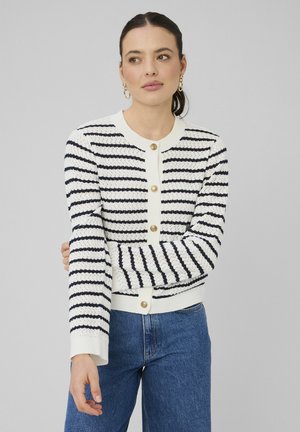 Femme portant un cardigan blanc texturé avec des rayures horizontales bleu marine et des boutons dorés, associé à un jean bleu, debout devant un fond uni.