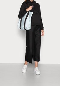 Černá mikina s logem, černé široké kalhoty, bílé tenisky a světle modrá taška s černými akcenty, držena v pravé ruce.