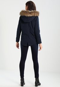Marineparka med pelsforet hætte, der har snøre i taljen, lange ærmer og slids i kanten. Bæres med mørke leggings og ankelstøvler.