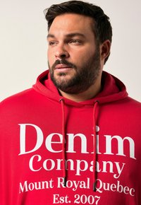 Sudadera roja con capucha que presenta un gran logo impreso en blanco "Denim Company", con texto más pequeño debajo. El diseño incluye una capucha con cordón y una textura de tela suave.