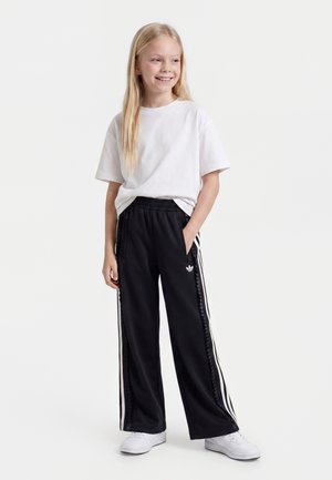 Fille souriante aux longs cheveux blonds porte un t-shirt blanc, un pantalon de survêtement noir Adidas avec des bandes blanches, et des baskets blanches, debout avec une main dans la poche.