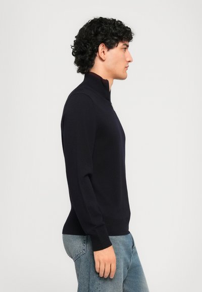 Man med lockigt hår som bär en svart zip-up-tröja och blå jeans stående i profil mot en enkel ljus bakgrund.