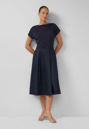 Kobieta stojąca przy ścianie, ubrana w granatowy krótki top z krótkim rękawem, pasującą spódnicę midi w linii A, srebrne kolczyki koła i srebrne sandały na obcasie.