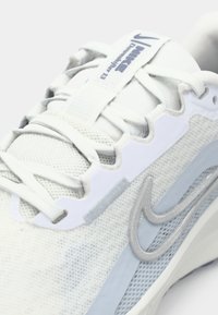 Gros plan d'une chaussure de course Nike Downshifter 13 blanche avec des lacets, un tissu en mesh et le logo swoosh gris sur le côté.