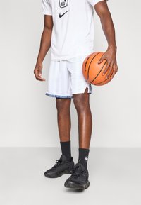 Shorts de baschet albe cu dungi albastre, având un model de tiv contrastant. Pantofi sport negri și un tricou alb cu logo-ul Nike NBA.