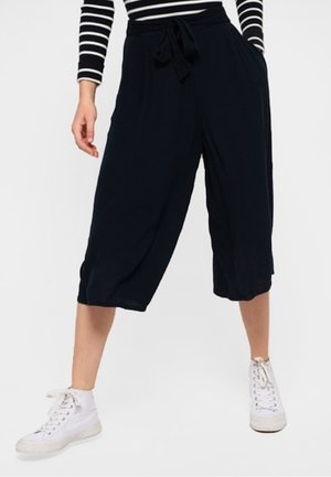 Woman wearing black wide-leg cropped pants with a tie waist and white high-top sneakers, standing against a plain background.