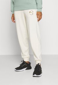 Light green sweatshirt with a kangaroo pocket, paired with cream sweatpants featuring a printed logo. Black athletic shoes with white soles.