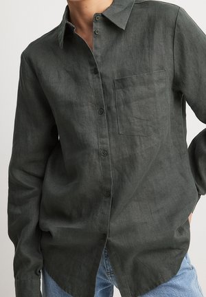 Mand iført en mørkegrøn, langærmet skjorte med brystlomme, kombineret med lyseblå jeans, mod en ensfarvet baggrund.