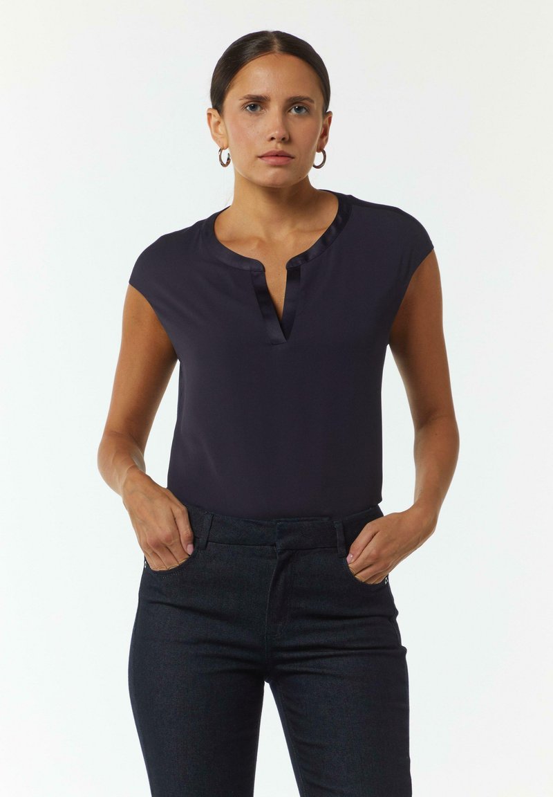 Blusa navy a maniche corte con scollatura a V e rifiniture in satin. Abbinata a pantaloni in denim scuro. Il tessuto appare liscio e aderente.