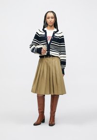 Ζακέτα με ναυτικό και λευκές ρίγες πάνω από λευκό πουκάμισο με κόκκινο κείμενο, συνδυασμένη με φούστα χακί με πτυχώσεις και καφέ μπότες μέχρι το γόνατο.