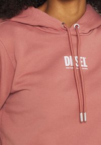 Felpa color rosa sporco con cappuccio regolabile, con logo e testo bianchi sul davanti. Tessuto morbido con una texture liscia.