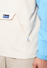 Hellbeige Pullover mit großer Fronttasche und blauen Ärmeln. Verfügt über ein Logo-Tag, elastische Bündchen und eine glatte Textur.