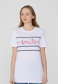 Camiseta de algodón blanca con la palabra "sonríe" en rojo, acompañada de dos franjas horizontales en azul marino en el pecho. Cuello redondo acanalado y mangas cortas.