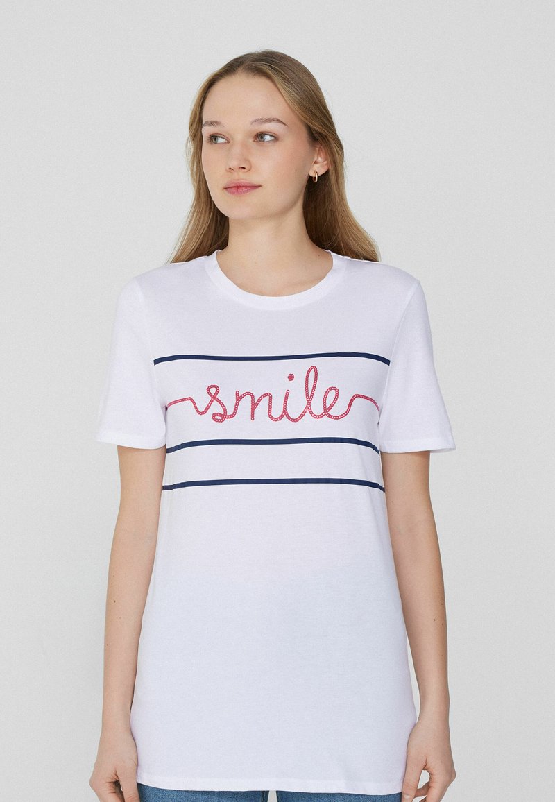Camiseta de algodón blanca con la palabra "sonríe" en rojo, acompañada de dos franjas horizontales en azul marino en el pecho. Cuello redondo acanalado y mangas cortas.