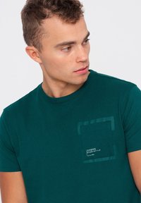 Zelené bavlněné tričko s kulatým výstřihem, s texturovanou kapsou a štítkem na levé straně. Hladká textura látky.