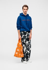 Μπλε φούτερ με κουκούλα με το κείμενο "marimekko", ασπρόμαυρο παντελόνι με σχέδια και μια πορτοκαλί τσάντα με μεγάλα λουλουδάτα μοτίβα. Άσπρα παπούτσια.