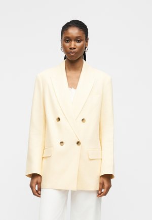 Vrouw met gevlochten haar, gouden oorringen, en een oversized crème dubbelgeknoopte blazer over witte broeken, staand tegen een effen achtergrond.