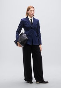 Μπλε σκούρο σακάκι με ρίγες και ένα κουμπί, σε συνδυασμό με μαύρο παντελόνι φαρδιάς γραμμής. Το μοντέλο κρατάει μια μαύρη τσάντα pouch, φοράει λευκό πουκάμισο και μαύρη γραβάτα.