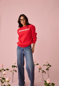 Femme portant un sweat-shirt rouge "Amour" et un jean bleu, debout parmi des roses roses dans des vases en verre, sur un fond rose.