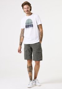 Camiseta blanca con un gráfico circular en azul y verde, combinada con pantalones cortos tipo cargo en verde oscuro. Presenta múltiples bolsillos y un corte relajado.