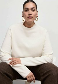 Top crema a maniche lunghe con un collo alto; texture del tessuto morbida. Il modello indossa grandi orecchini a cerchio dorati e siede su una sedia bianca. Pantaloni marroni.