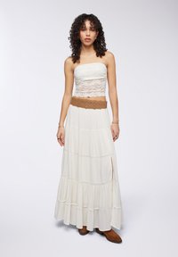 Top corto senza spalline in pizzo bianco abbinato a una gonna maxi fluida color crema con strati a balze e una cintura decorativa marrone.