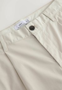 Beige bukser med almindelig tilspidset pasform, lukkes med knap, har bærestropper og synligt NEXT-mærke inde i linningen.