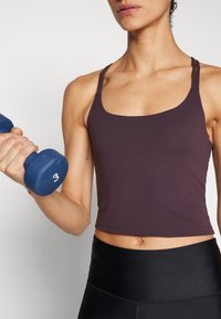 Lilla atletisk crop top med tynde stropper og tætsiddende design, parret med sorte højtaljede leggings. Holder en blå håndvægt mærket "3."