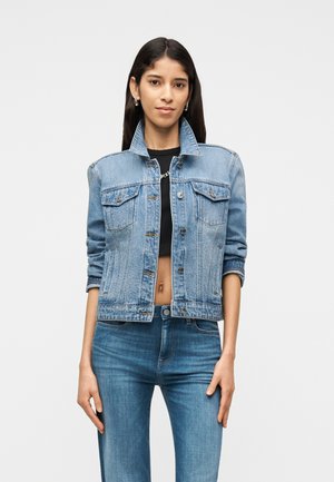 Femme portant une veste en jean bleu clair sur un haut court noir et un jean bleu, debout devant un fond clair uni.