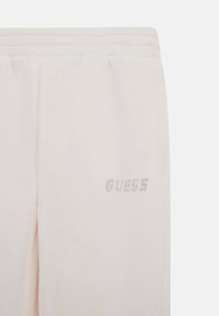 Pantalones jogger de color beige claro con una cinturilla acanalada, textura de tela suave y el logo "GUESS" en strass plateados en la parte inferior de la pierna.