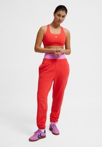 Rode sportbeha met racerback-ontwerp, gecombineerd met loszittende rode trainingsbroeken met een roze tailleband en paarse schoenen met blauwe accenten.