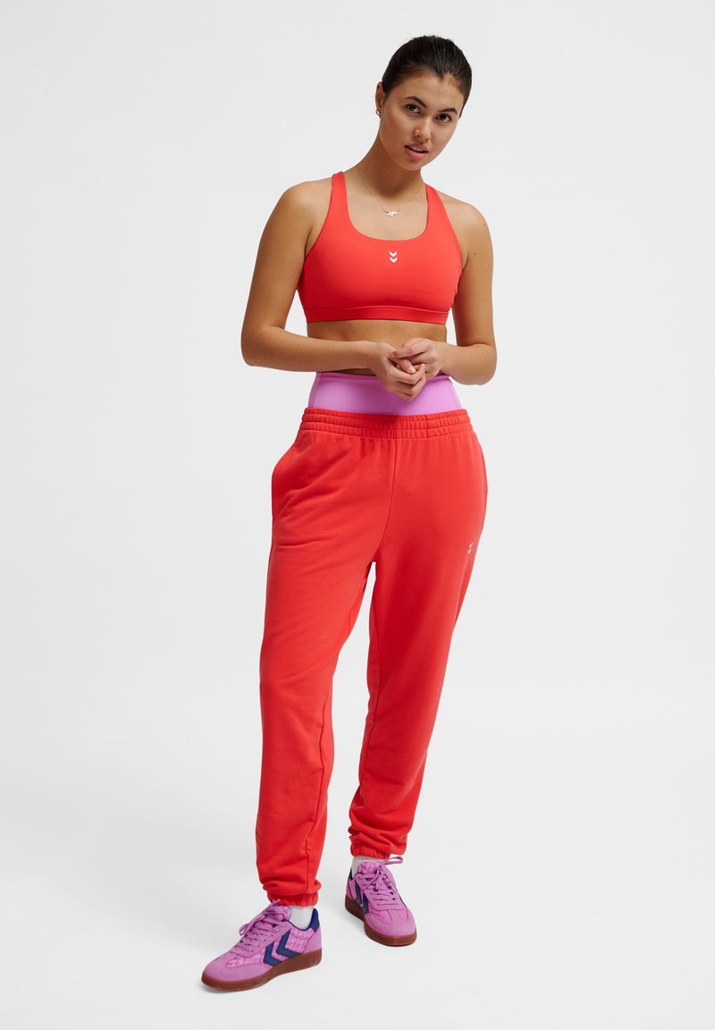 Rode sportbeha met racerback-ontwerp, gecombineerd met loszittende rode trainingsbroeken met een roze tailleband en paarse schoenen met blauwe accenten.