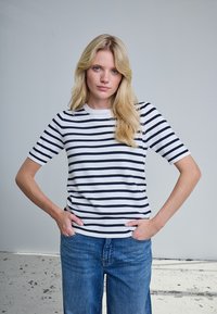 Femme blonde aux cheveux lâchés, debout les mains dans les poches, portant un t-shirt à manches courtes blanc et noir à rayures et un jean bleu.