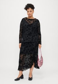 Juoda gėlėta nėriniuotė palaidinė su ilgomis rankovėmis, dėvima ant juodos sijono. Palaidinė turi garbanotą apatinį kraštą, o ji derinama su rožine rankine ant peties ir juodais batais.