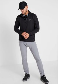 Férfi fekete sapkában, fekete Calvin Klein negyedcipzáras pulóverben, világosszürke nadrágban és fekete tornacipőben, összekulcsolt kézzel áll.